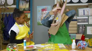 Een foto van Railey in gele schort en Rosalie in groene schort achter een tafel. Rosalie heeft een lichtbruine papieren zak vast met een gat voorop waar ze haar hand doorheen steekt en erboven is een geknutselde paashaaskop te zien. Railey kijkt ernaar.