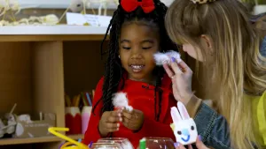 Een foto van Geneely die een rode jurk aanheeft en een rode strik op haar hoofd. Ze heeft een wit veertje in haar hand. Rechts heeft Rosalie een wit veertje in haar rechterhand en in haar linkerhand heeft ze een witte paashaaskop met blauwe neus en mond. 