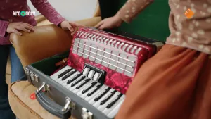 Een foto van een rood met zwarte accordeon op een stoel in een zwarte koffer. De accordeon heeft toetsen zoals op een piano maar ook een opgevouwen zak in het midden, die je tijdens het spelen in de breedte uittrekt en daarna weer in elkaar drukt.