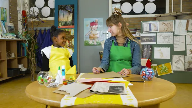 Een foto van een meisje in een gele schort en Rosalie in een groene schort die achter een tafel zitten en naar elkaar kijken. Op de tafel liggen papieren en kartonnen vellen, een geel met wit kleed en een krant. Ook staan er knutselspullen en lijm.