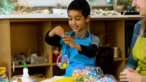 Een foto van Ahmed die achter een tafel staat en een veelkleurige hanger met paaskuikens aan een blauw lint vastheeft. De hanger bestaat uit een cirkel van allemaal gekleurde stukjes en drie paaskuikentjes erop in roze, groen en geel. Rosalie zit rechts. 