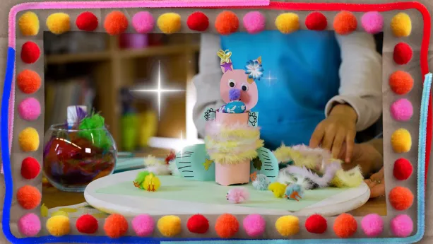 Een foto van een uit een wc-rol gemaakte paashaas die roze is, een paarse neus heeft met een vlinder, sterretje en bloem op de oren. In de rol zitten paaseitjes en ervoor liggen zes paaskuikentjes. Om de foto staan knutselbolletjes en knutselstaafjes.