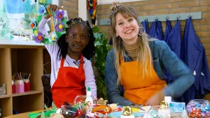 Een foto van Rosalie met oranje schort en Djah-Relly met rode schort die aan een tafel vol knutselartikelen zitten. Djah-Relly houdt een bloemenkrans omhoog met blauwe en paarse bloemen met rode en gele knoppen. De bloemenkrans is gemaakt van eierdozen.
