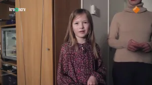 Een foto met Elana in het midden. Elana draagt een rode jurk met bloemetjes erop van een lichter rood en witte blaadjes. Naast Elana is links een lichtbruine kast met een deur en planken te zien. Rechts is haar moeder tot net boven haar kin te zien.