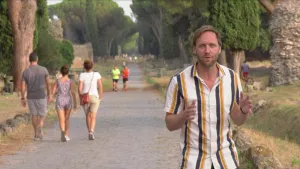 Presentator op een oude Romeinse weg waarop toeristen wandelen.