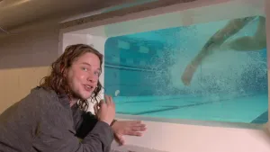 Presentator staat bij raam met zicht op zwembad, onderwater, been van zwemmer die vele luchtbelletjes veroorzaakt.