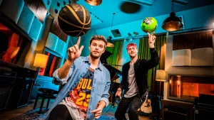 Een foto van Buddy in een basketbalshirt van de ploeg Chicago Bulls en meester Willem met rode zweetband. Buddy houdt een basketbal, zwart met gouden lijnen, omhoog op één vinger, meester Willem een voetbal met groene en zwarte vlakken op twee vingers.