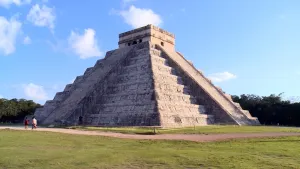 Een foto van de Piramide van Kukulcán in Mexico, een tempel gebouwd door de Maya's. Twee trappen leiden naar boven aan twee zijden van de piramide. De stenen zijn grijsachtig van kleur en deels lichtoranje verlicht door de zon. Er lopen drie mensen voor.