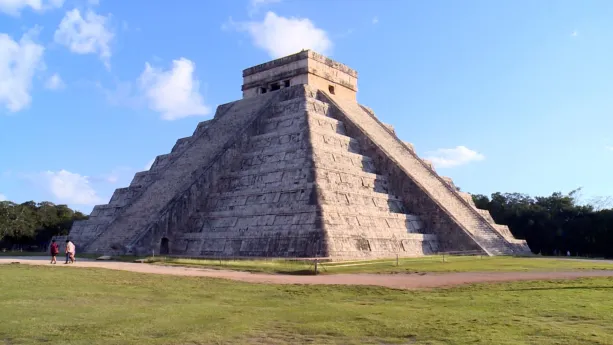 Een foto van de Piramide van Kukulcán in Mexico, een tempel gebouwd door de Maya's. Twee trappen leiden naar boven aan twee zijden van de piramide. De stenen zijn grijsachtig van kleur en deels lichtoranje verlicht door de zon. Er lopen drie mensen voor.
