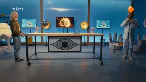 Een foto van presentator Janouk in de studio van Brainstorm. Ze staat links van een tafel. Aan de rechterkant staat een man met een baard. Hij is met een statief bezig.