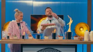 Een foto van presentator Janouk. Ze staat in een Tv-studio naast Job. Ze staan achter een tafel met daarop enkele potjes waar vlammen uit komen. Job houdt een tray met eieren in een hand en een los ei in zijn andere hand. 