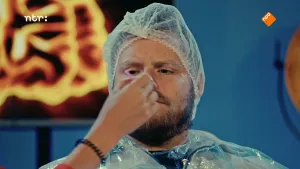 Een foto van een man met een baard. Hij heeft een soort regenjas aan, en een haarnetje op zijn hoofd. Iemand houdt iets voor zijn mond. Hij kijkt ernaar.
