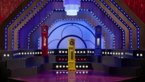 Foto van een soort podiumachtige ruimte met verlichting waarin drie klein nagemaakte staande klokken staan, een blauwe, gele, rode.