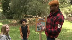 Een foto van boswachter Tim. Hij staat in de natuur en heeft een Beestenbrigade bingo bord geplaatst. Hij wijst naar naar bord. Naast hem staat een meisje en een jongen. 