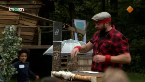 Een foto van boswachter Tim die buiten achter een tafel staat. Op die tafel staat een glazen bak met eikels. Hij stopt zijn hand daarin en is ook wat aan het vertellen. Op de achtergrond zie je een houten hut en bomen.