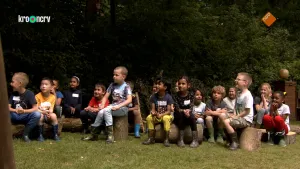 Een foto van ongeveer twintig kinderen die buiten zitten, op boomstammen. Ze kijken allemaal dezelfde kant op. De meeste van hen kijken blij. Op de achtergrond zie je bomen.