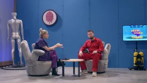 Een foto van presentator Janouk en prof die beiden in een stoel zitten. Prof heeft een rood pak aan en kijkt naar de camera. Janouk wijst met beide handen naar prof. Ze zitten op een grijze stoel waarvan de rugleuning en armleuningen op vingers lijken.