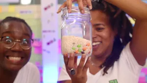 Een foto van de twee presentatoren Tirsa en Eva. Tirsa kijkt enthousiast de camera in. Eva kijkt blij naar de glazen pot in haar hand. In de pot zit rijst met gekleurde bolletjes. 