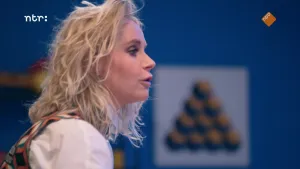 Een foto van presentator Janouk. Achter haar is een muur te zien met een poster. Op de poster staat een driehoekige vorm. Janouk is van opzij te zien, aan de linkerkant van de foto. Ze praat tegen iemand die niet te zien is.