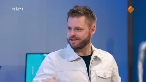 Een foto van een man in de studio. Achter hem is een stukje van een computerscherm te zien en een muur.