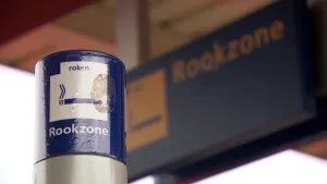 Een foto van een stalen paal waar aan de zijkant de woorden "roken" en "Rookzone" te lezen zijn met het plaatje van een sigaret erop. Op de achtergrond is ook een groot blauw bord zichtbaar met dezelfde teks en een plaatje van een sigaret erop.
