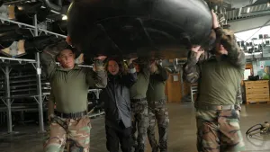 Een foto van presentator Pascal die samen met vijf mariniers een zwarte rubberboot draagt in een ruimte van een gebouw. Ze dragen de boot boven hun hoofd. De mariniers hebben speciale marinierskleding aan, een groene blouse en een broek in legerkleuren.