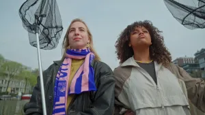 Een foto van presentator Quinty die rechts in beeld is en een vrouw links op de foto die met een jas aan buiten staan. Links is nog net te zien dat ze aan de waterkant staan in een woonwijk. Ze hebben allebei een zwart visnet aan een lange stok vast.