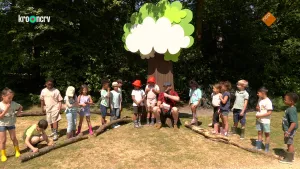 Een foto van Boswachter Tim die op een boomstronk zit voor een platte nepboom met groene bladeren en een witte wolk ervoor. Hij leest een vraag voor in een microfoon aan twee kinderen met een rood hoedje. Andere kinderen staan ernaast achter dikke takken.