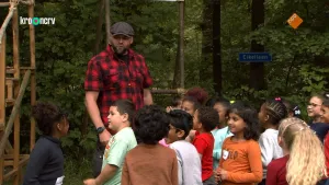 Een foto van Boswachter Tim die in een rood met zwart geruite blouse en een zwarte pet voor een groep kinderen staat in een open plek in een bos. Rechts achter de kinderen hangt een blauw straatnaambord met de naam "Eikellaan" erop op een boom.  
