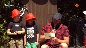 Een foto van Boswachter Tim die in een rood met zwart geruite blouse en een zwarte pet met grijze strepen buiten zit. Hij heeft een kaartje vast met vragen en leest een vraag voor in een microfoon. Twee kinderen met een rood hoedje op staan links van Tim.