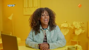 Een foto van Quinty die in een kamer zit waarin alles geel gekleurd is. Ze zit achter een bureau met een laptop, een hoofdtelefoon en een beker erop. Erachter staat een plant op een kruk en hangt een schilderij met drie ijsjes erop.