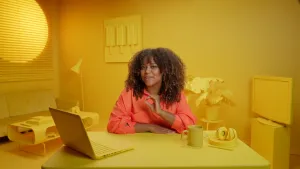 Een foto van Quinty die in een kamer zit waarin alles geel gekleurd is. Ze zit achter een bureau met een laptop, een hoofdtelefoon en een beker erop. Op de achtergrond staan een bank, een tafel, hangt een schilderij met drie ijsjes erop en staat een tv.