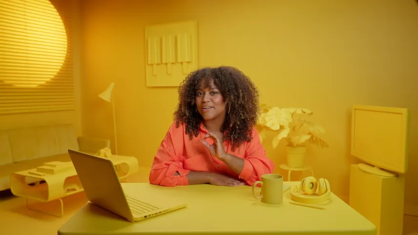 Een foto van Quinty die in een kamer zit waarin alles geel gekleurd is. Ze zit achter een bureau met een laptop, een hoofdtelefoon en een beker erop. Op de achtergrond staan een bank, een tafel, hangt een schilderij met drie ijsjes erop en staat een tv.