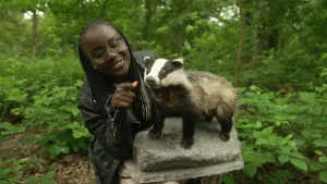 Een foto van Tirsa die in een omgeving met groene planten achter een stapel stenen staat met daarop een opgezette das. De das heeft een zwarte en witte vacht met zwart-wit gestreepte kop, zwarte snuit en zwarte nagels. Tirsa wijst naar zijn snuit. 
