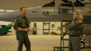 Een foto van een piloot in een pilotenpak links en Janouk in een pilotenpak rechts. Janouk staat naast een trap naar de open cockpit van een straaljager. Een straaljager is een heel snel gevechtsvliegtuig, waarmee de lucht goed bewaakt kan worden. 