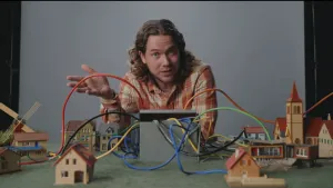 Een foto van Pascal die achter een tafel staat met een open bak, waar allemaal gekleurde kabels uitkomen. Die kabels gaan allemaal naar modellen van huisjes, een molen en een kerk op de tafel toe. Dit laat een stukje van de werking van internet zien.