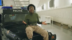 Een foto van Tirsa op de motorkap van een zwarte politieauto. Op de auto staat aan de zijkant het logo van de politie en boven het voorraam met zwart gaas ervoor staat "Politie". Op de auto staan blauwe en witte lichten voor een rond oranje zwaailicht.