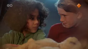 Een foto van Saïd en Anna die achter een tafel staan. Ze kijken naar elkaar. Voor hen is de onscherpe vorm van een lichtbruine teddybeer te zien die op zijn rug ligt. Saïd draagt een groene trui met touwtjes eraan en een rits en Anna een rode trui.