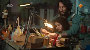 Een foto van Saïd die voor zijn vader achter een werktafel zit. Op de tafel staan potjes met kralen. De vader is bezig een kralenketting te maken en heeft een deel van de ketting, die niet te zien is, vast. Een tangetje steekt achter zijn hand omhoog.