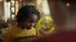 Een foto van een kind in een donkergeel vest die naar beneden kijkt in een zwarte kooi. Op de kooi is onscherp een geel doorzichtig plastic, loopwiel voor een hamster te zien. Rechts vooraan zijn de rug en nek van een andere persoon te zien.