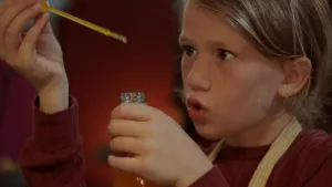 Een foto van Anna die een doorzichtig staafje met geel vocht erin, een pipet, vastheeft in haar rechterhand en een glazen flesje in haar linkerhand. Ze kijkt met aandacht naar de pipet. Ze wil een druppel van de pipet in het flesje laten druppelen.