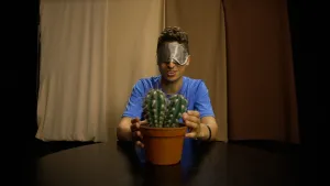 Een foto van presentator Nizar die achter een tafel zit. Nizar heeft een oogmasker op en voelt met zijn handen aan de stekels van een cactus die op de tafel staat. Achter Nizar hangt een lichtbruin gordijn.