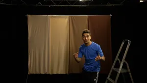 Een foto van presentator Nizar in een donkere ruimte. Naast Nizar staat een trap op de grond. Achter Nizar hangt een lichtbruine gordijn aan een stang.