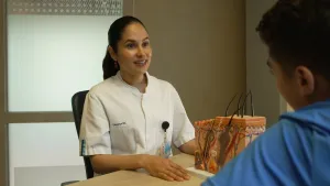 Een foto van Petra achter een bureau met Nizar. Voor Petra staat een plastic voorbeeld van de menselijke huid. Er zitten haren op het voorbeeld en bloedvaten en haarzakjes zijn zichtbaar aan de zijkant. Petra is een dermatoloog, dat is een huiddokter. 