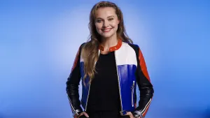 Een foto van zangeres en actrice Vajèn van den Bosch. Ze lacht naar de camera en heeft een rood-wit-blauwe leren jas aan met deels zwarte mouwen. Ze heeft haar handen in haar zakken en haar duim op haar riem.  Ze staat voor een blauwe achtergrond.