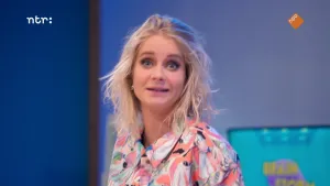 Een foto van Janouk die met licht opgetrokken wenkbrauwen en grote ogen naar de camera kijkt. Ze draagt een witte blouse met kleurige patronen in roze, groen en blauw met kleine zwarte stukjes erdoorheen.