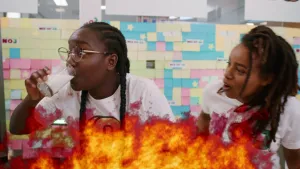 Een foto van presentatoren Tirsa en Eva. Voor de foto is een brandend vuur zichtbaar. Tirsa neemt een slok van een glas melk terwijl Eva bezorgd toekijkt. Tirsa neemt een grote slok melk, omdat ze net een hap van de hele hete peper Habanero heeft genomen.