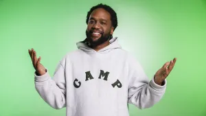 Een foto van de Nederlandse rapper en zanger Glen Faria die lacht naar de camera met zijn handen met de handpalmen naar boven in de lucht. Hij heeft een witte capuchontrui aan met in het zwart de letters "CAMP" erop. De achtergrond van de foto is groen.