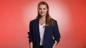 Een foto van zangeres en actrice Vajèn van den Bosch. Ze lacht naar de camera en heeft een donkerblauw pakjasje met een witte blouse eronder aan. Ze staat voor een rode achtergrond.