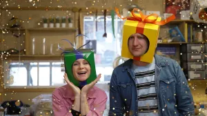 Een foto van Janouk en Maarten Bel met een pakje om hun hoofd. Janouk een groen pakje met paarse strik, Maarten een geel pakje met oranje strik. Janouk houdt haar armen tegen elkaar met de handpalmen omhoog en kijkt blij. Maarten glimlacht. 